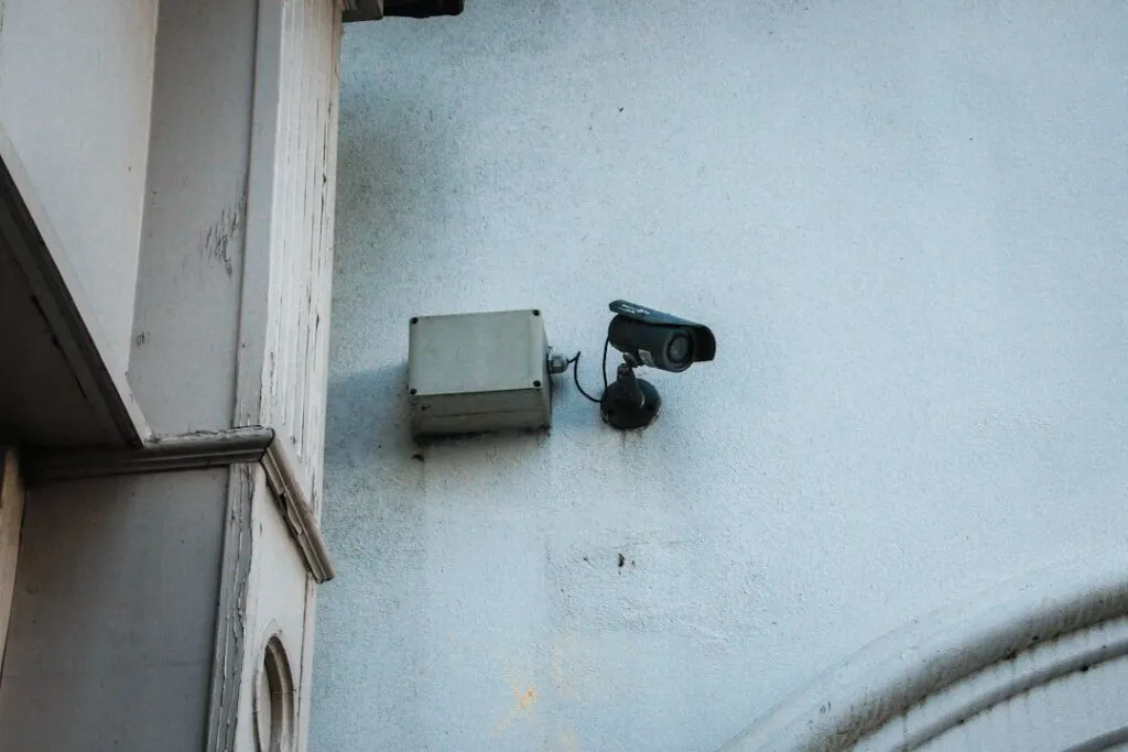 Bug Sweep Services A security camera for surveillance and a gray utility box are installed on a white wall, providing reliable building monitoring. Infidelity Investigator
