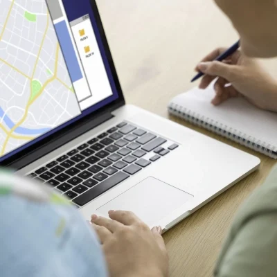 Two private investigators work at a table, one analyzing a map on a laptop while the other takes detailed notes in a notebook.