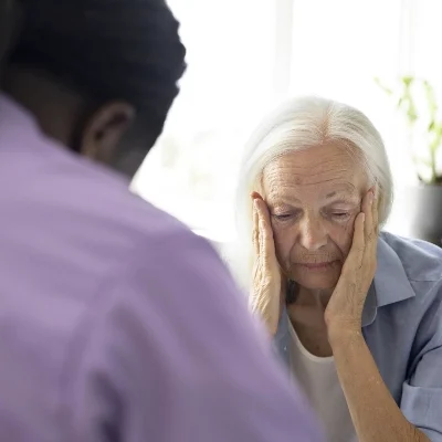 A worried elderly woman sits with her hands on her face, possibly seeking help from a private investigator in a bright office.