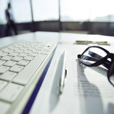A computer keyboard, pen, document, and eyeglasses on a desk in a bright office; a figure stands in the background—private investigator workspace.