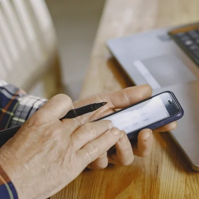 Private investigator using a smartphone with a stylus next to a laptop on a wooden table, researching confidential information.
