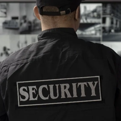 A professional in a SECURITY jacket and cap monitors several surveillance screens, highlighting expert surveillance and investigation skills.