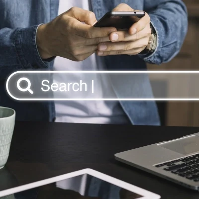 Private investigator researching on a smartphone near laptop and tablet, with digital search bar overlay symbolizing online investigation.