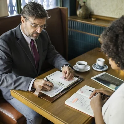Two people at a table with notebooks and coffee, discussing investigation plans; one writes while the other gestures to a planner.