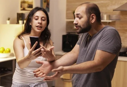 A woman displays a phone to a man in a kitchen, possibly revealing evidence during an intense conversation with a private investigator.