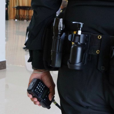 A professional private investigator in a black uniform holds a two-way radio, with a duty belt featuring flashlight and investigation tools.