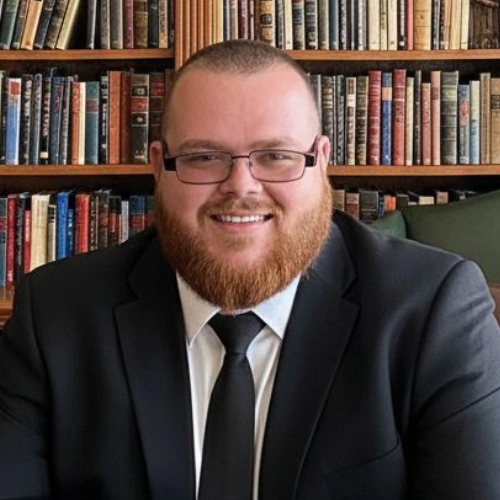A professional private investigator with a red beard and glasses, in a black suit and tie, sits before shelves of books.