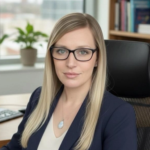A professional female private investigator with long blonde hair and glasses works at her office desk, shelves and plant behind her.