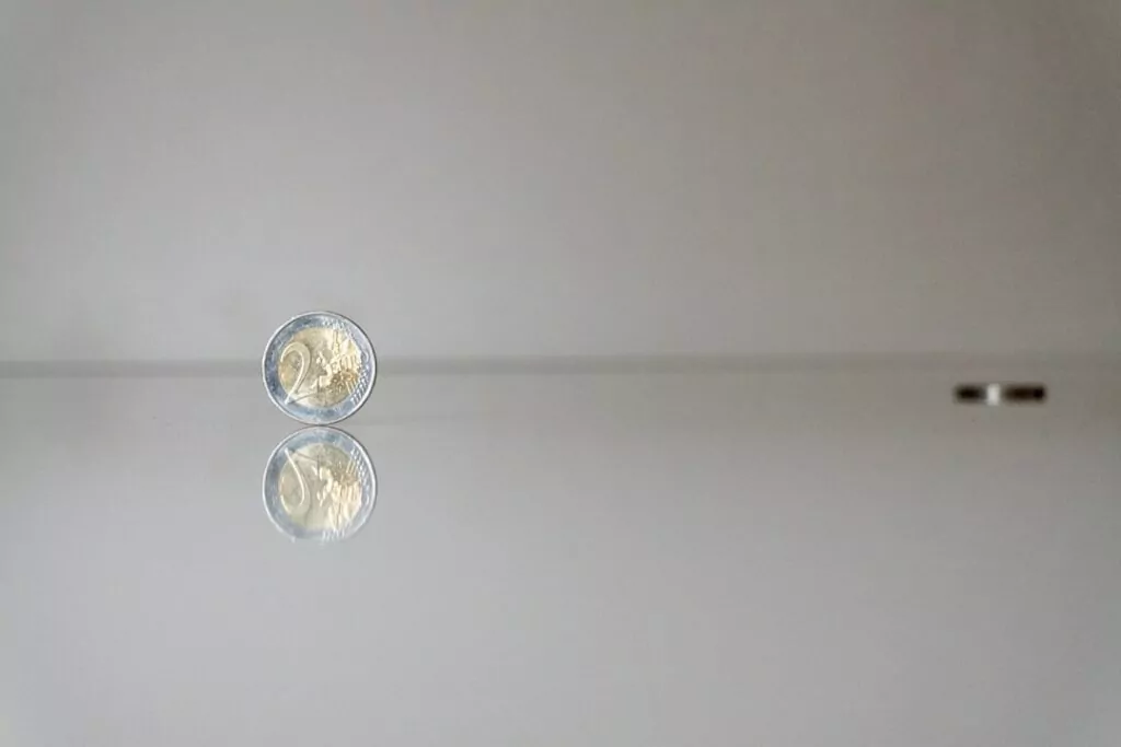 A two-euro coin stands upright on a reflective surface, symbolizing clarity and transparency, with a blurred coin in the background.