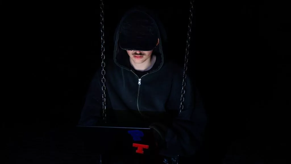 A private investigator in a dark hoodie and cap sits on a swing at night, working on a laptop, chains hanging on each side.