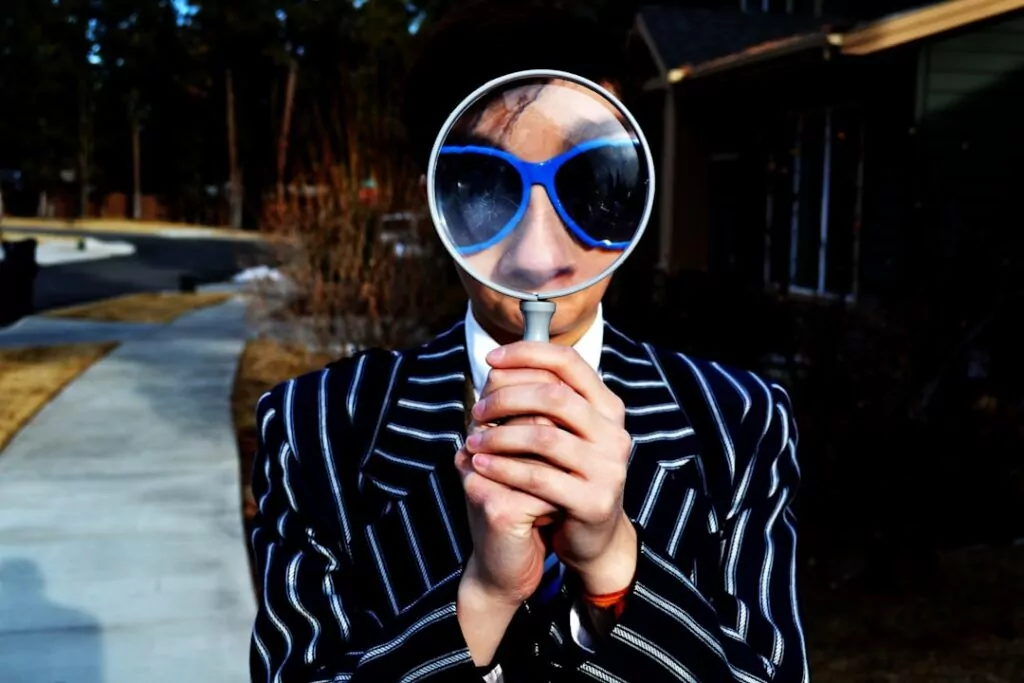 Professional private investigator in a striped suit and blue sunglasses uses magnifying glass, searching evidence outdoors by sidewalk.