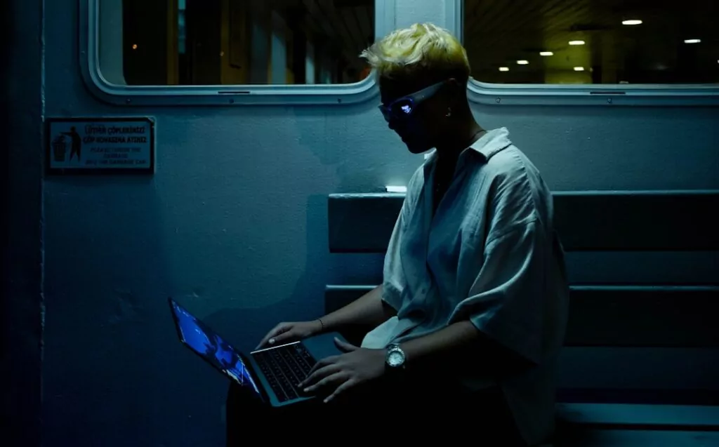 Private investigator with short blond hair and glasses researches on a laptop in low light; informative sign visible behind them.